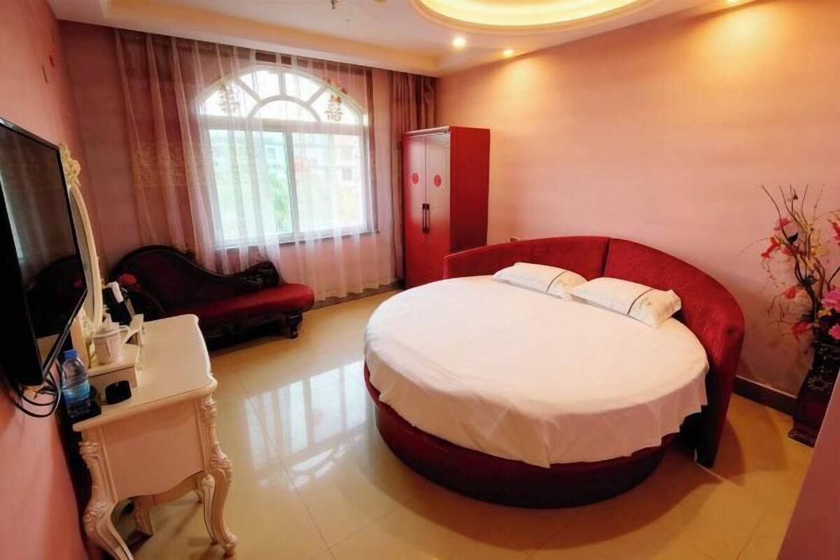 Hotelkamer met een groot rond bed, een rode garderobe, een klein nachtkastje met een lamp en een groot raam met gordijnen.