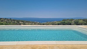 Una piscina rectangular con agua azul cristalina, rodeada por una terraza de hormigón y con vistas a un paisaje costero con colinas y al mar.