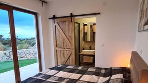 A bedroom with a large bed, a bedside table with a lamp, a sliding barn door, a bathroom with a sink, and a view of a green landscape through a large window. - Villa SOL Insulam (Novalja)