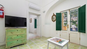 A room with a television, a green dresser, a table with a magazine, green shutters, and a view of buildings through the door. - Casa Bijou (Positano)