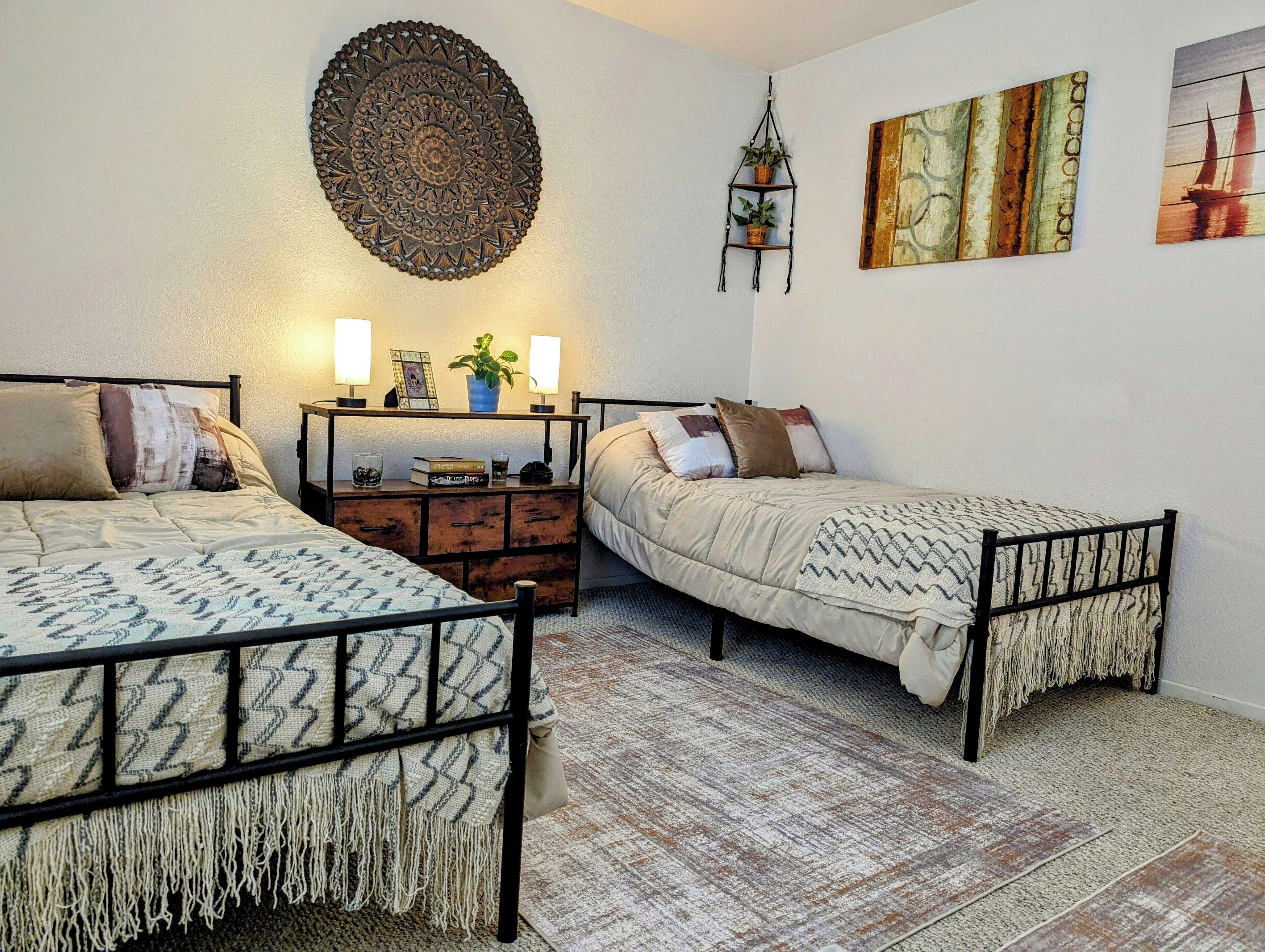 A bedroom with two beds, a bedside table with lamps, a wall hanging, and a painting.