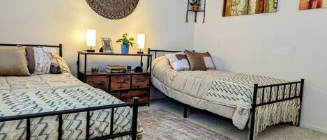 A bedroom with two beds, a bedside table with lamps, a wall hanging, and a painting.
