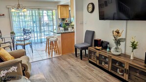 Un salón amplio con televisor, asientos cómodos, una zona de comedor con mesa de cristal y una cocina al fondo.