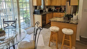 Una cocina con gabinetes de madera, encimera de granito, refrigerador de acero inoxidable y un comedor con mesa de vidrio y taburetes.