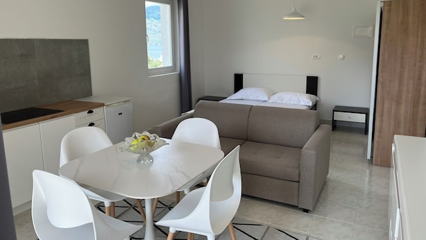 Un espace de vie moderne et compact, comprenant un coin cuisine, un espace repas avec une table et des chaises blanches, un canapé-lit et une grande fenêtre offrant une vue sur l’extérieur.