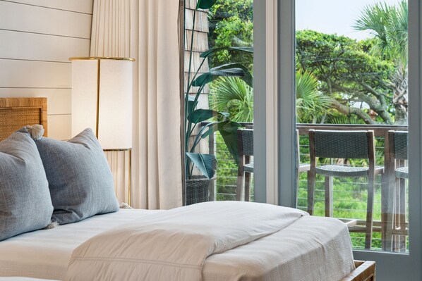 Un dormitorio con una cama grande, una mesita de noche y vistas a un balcón con sillas y vegetación.