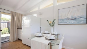 Uma sala de jantar moderna com uma mesa de vidro posta para quatro pessoas, cadeiras brancas e um quadro com barcos à vela na parede.