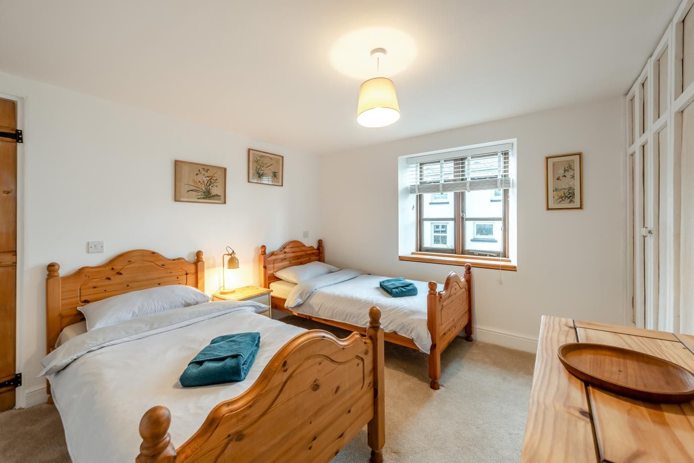 A bedroom with two wooden beds, a window with blinds, and a bedside table with a lamp.