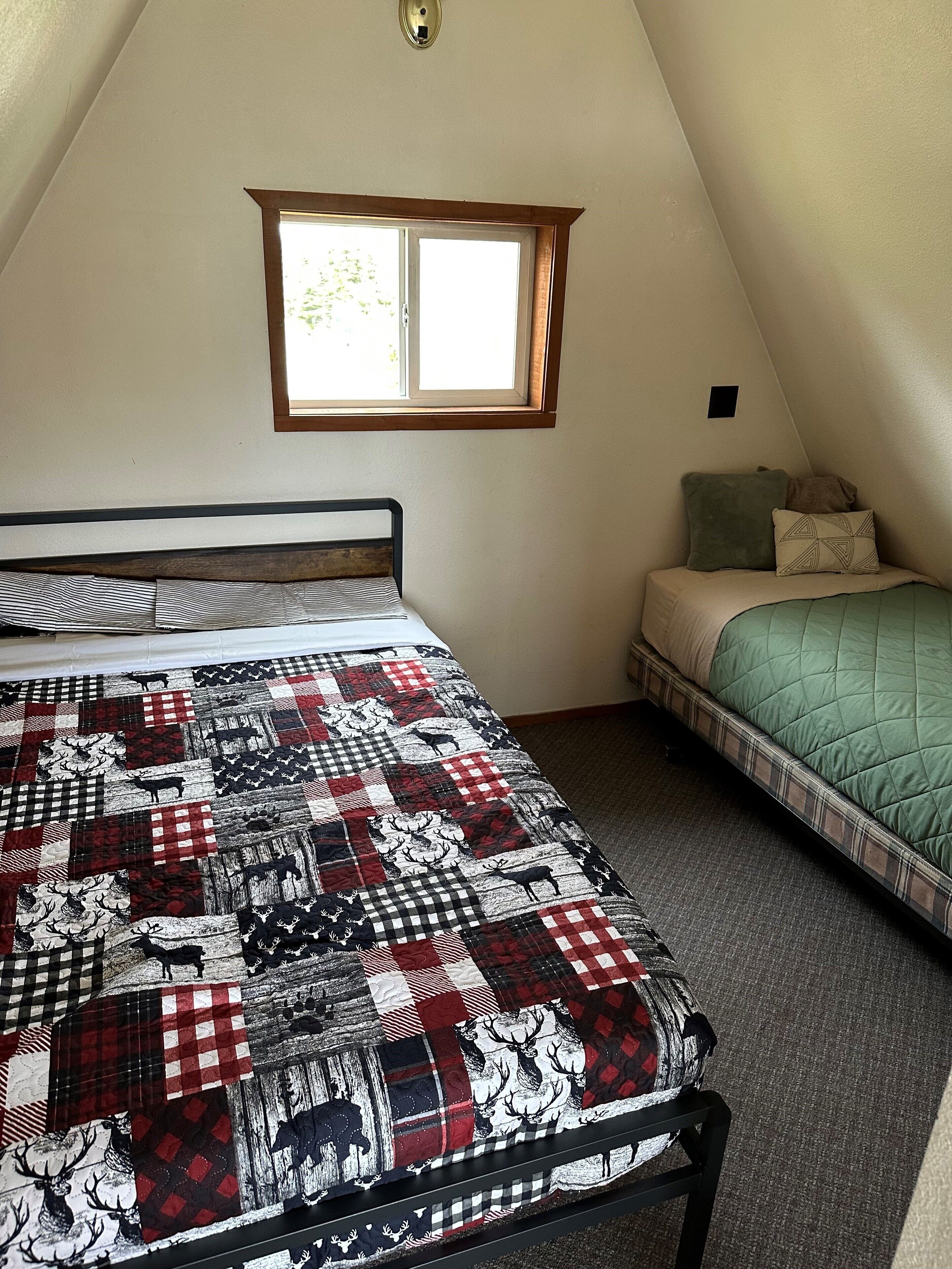 A bedroom with a large bed covered in a red and black checkered quilt, a smaller bed with a green quilt, a window with a wooden frame, and a small wall-mounted light fixture.