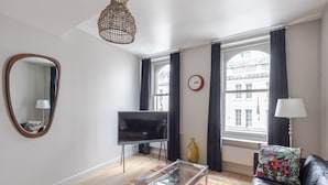 A modern living room with a glass coffee table, a leather sofa, a flat-screen TV, a large mirror, and a window with curtains. - Immaculate 2-bed Apartment in Prime Central London (London)