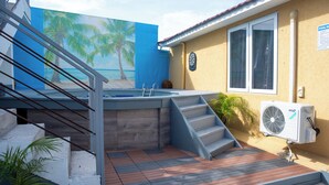 Una zona de piscina con una escalera que lleva a una terraza, un ventilador y un mural de palmeras y una playa.