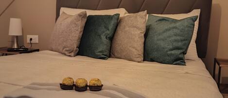 Un lit bien fait, avec une serviette blanche pliée et trois chocolats emballés, une table de chevet avec une lampe et une figurine décorative posée sur la tête de lit.