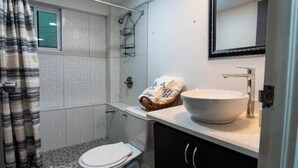 Une petite salle de bain avec un lavabo blanc, des toilettes et un espace douche. On y trouve un porte-serviettes et un sac de pain sur le comptoir.