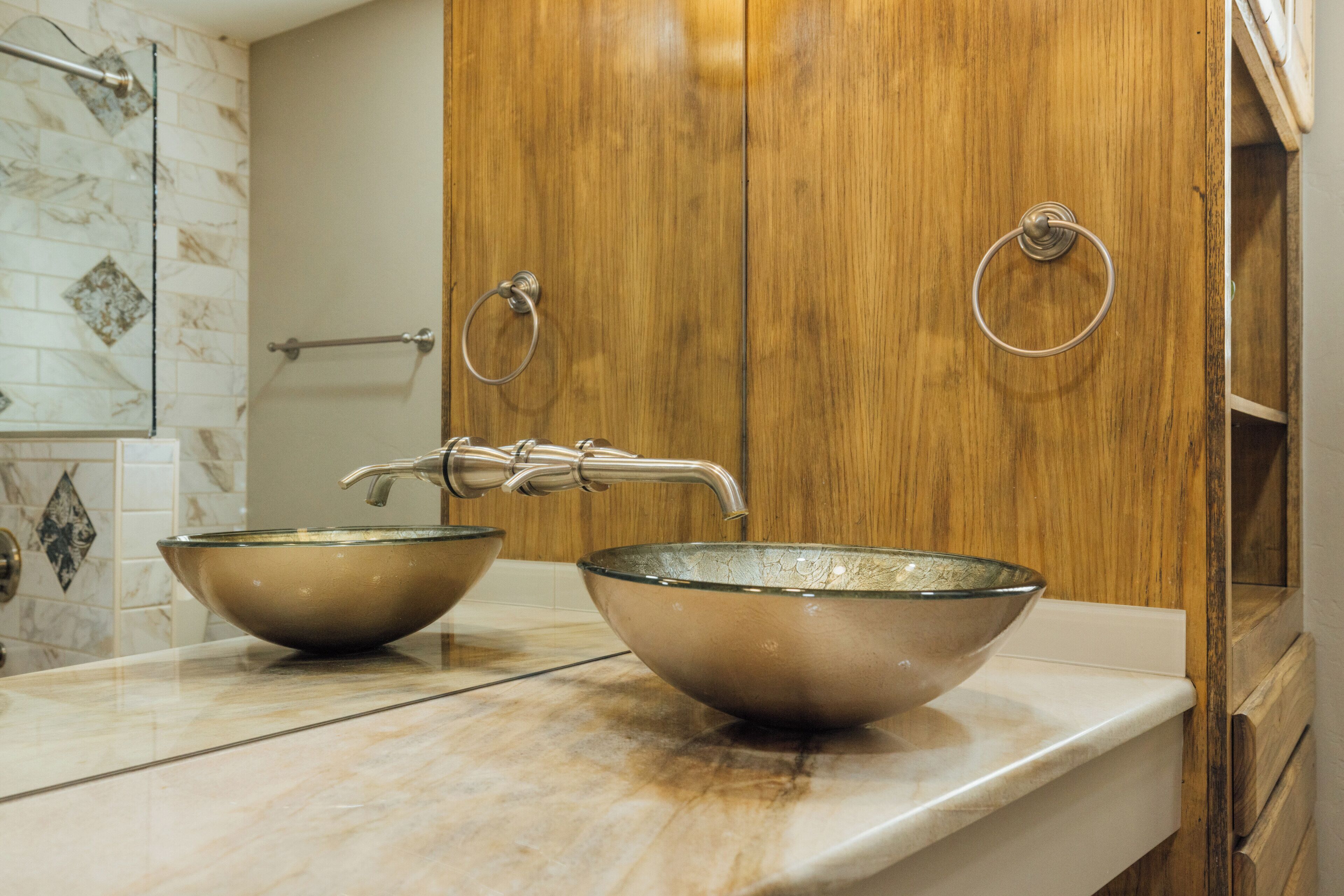 A bathroom with two glass bowls on a marble countertop, wooden cabinets, and a towel rack.