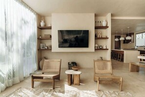 A living room with two wooden chairs, a coffee table, a mounted painting, and a kitchen area in the background.