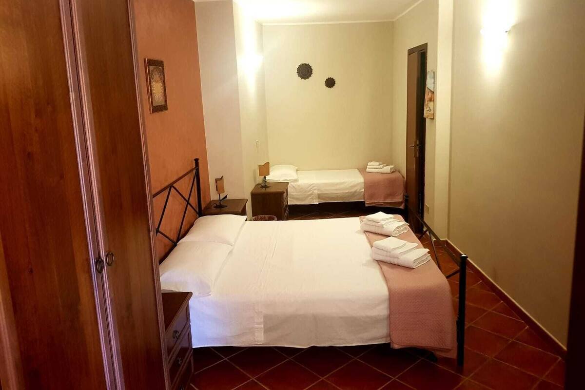 A hotel room with two beds, a wooden wardrobe, a small bedside table, and a wall-mounted light fixture.