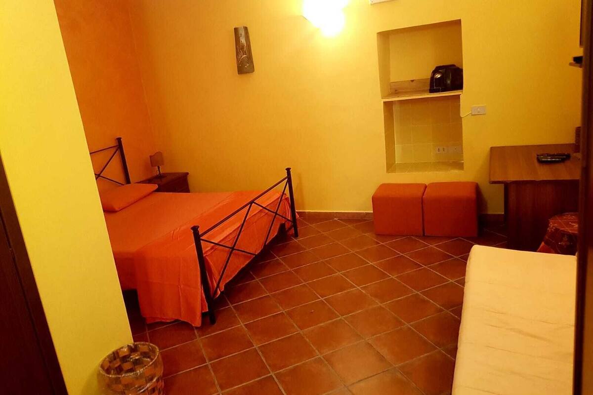 A small, brightly lit room with a bed, a nightstand, a shelf with a kettle, and a tiled floor.