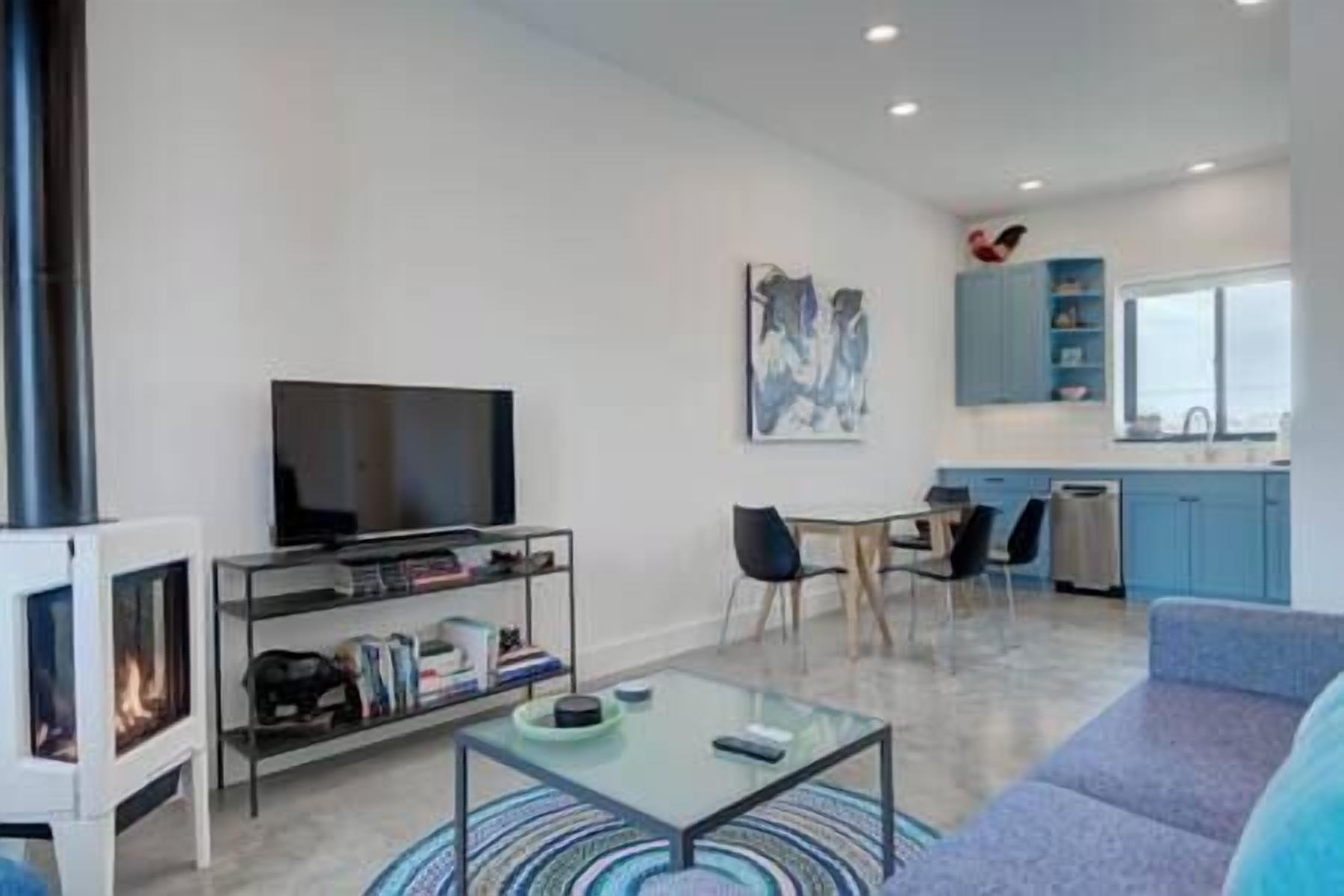 A modern living room with a flat-screen TV, a glass coffee table, and a sofa. There is a bookshelf with books and a fireplace. The kitchen in the background has blue cabinets and a window.