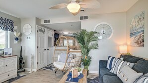 Ein Wohnzimmer mit grauem Sofa, einem Korbstuhl, einem Holztisch mit einem Buch und einer großen Wanduhr.