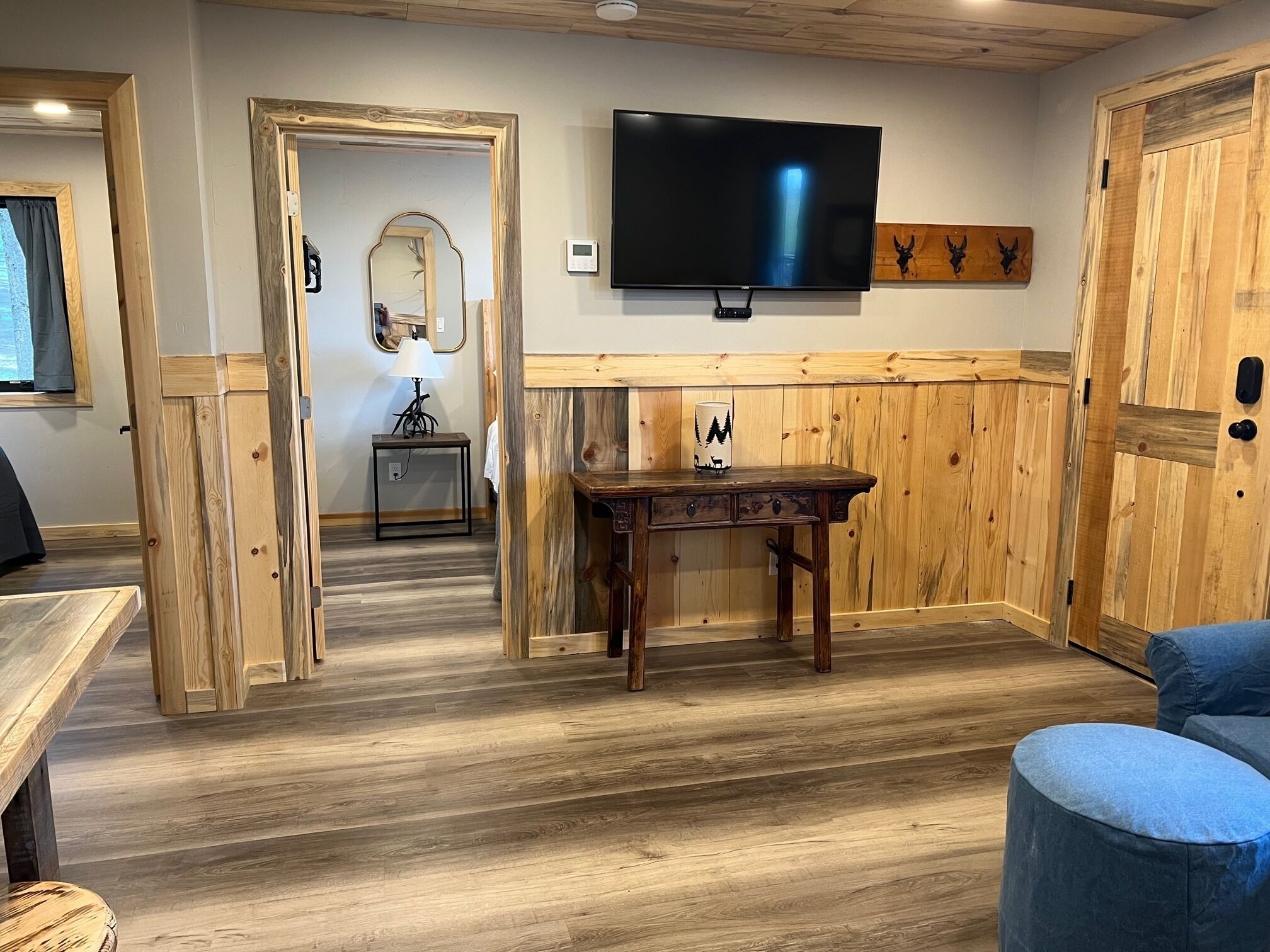A room with a wooden floor, a TV mounted on the wall, a table with a lamp, and a chair.