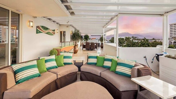 A covered outdoor seating area with a sofa and table, a view of the cityscape, and a sunset. - The Dreamscape: Sea View Serenity on the Esplanade (Cairns)