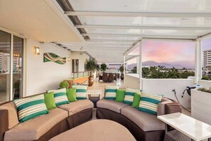 Un espace extérieur aménagé avec un coin salon couvert, comprenant un canapé et une table, offrant une vue sur le paysage urbain et un coucher de soleil.