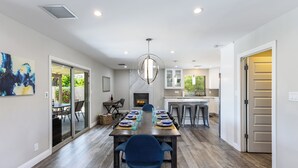 Un espace de repas moderne avec une longue table dressée pour six personnes, une cheminée et une cuisine en arrière-plan.