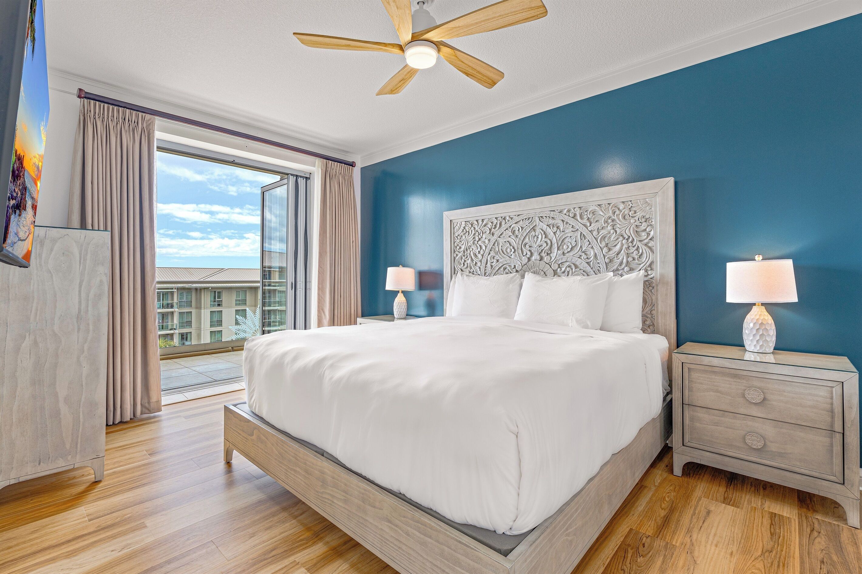 A hotel room with a large bed, a ceiling fan, a nightstand with a lamp, and a view of the city through the window.