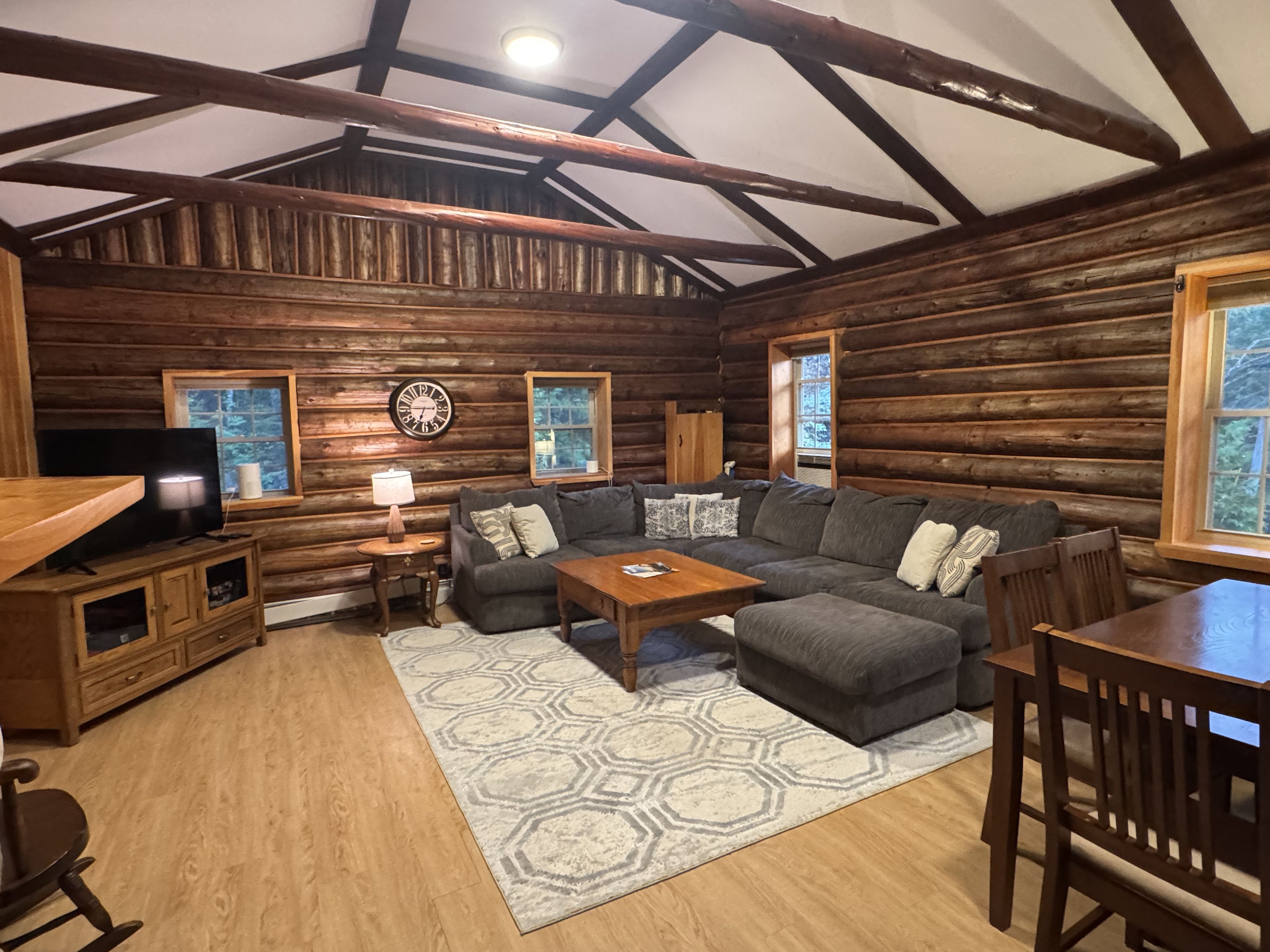 A log cabin interior with a sectional sofa, a television, a wooden table, and a patterned rug.