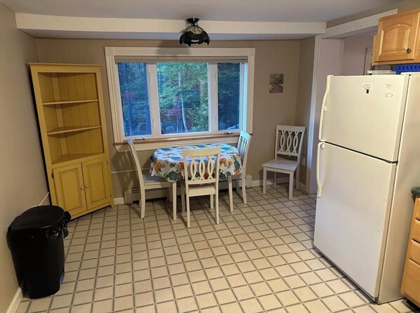 Een kleine keuken met een raam, een tafel met stoelen, een koelkast en een vuilnisbak.