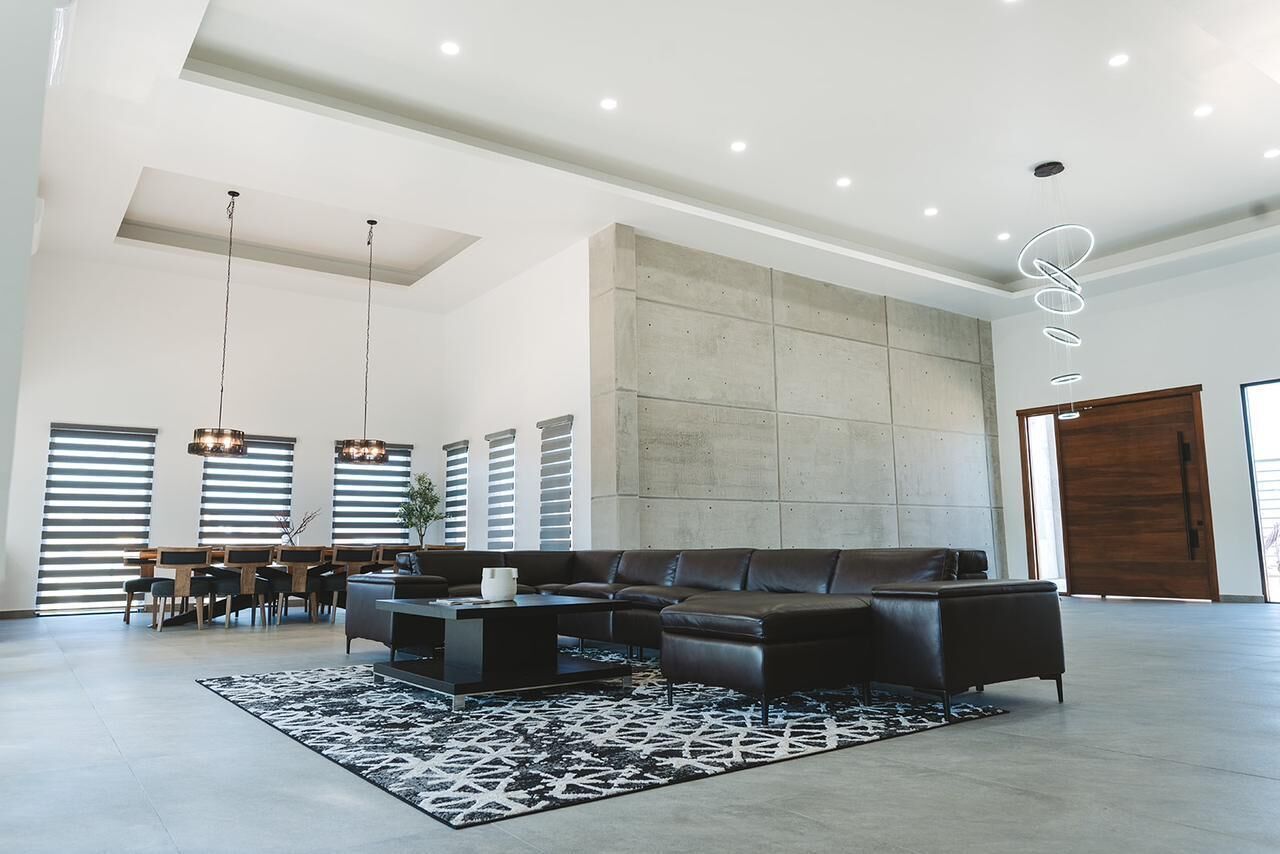 A spacious living room with a sectional sofa, a coffee table, and a patterned rug. There are pendant lights, a dining area with a table and chairs, and a large window with blinds.