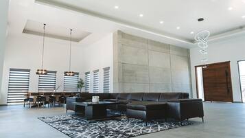 A spacious living room with a sectional sofa, a coffee table, and a patterned rug. There are pendant lights, a dining area with a table and chairs, and a large window with blinds.