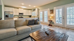 A modern kitchen with white cabinets, a refrigerator, and a microwave. There is a beige sofa with pillows and a wooden coffee table with a remote control.