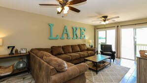 Un salon avec un grand panneau indiquant « LAKE », un canapé d’angle marron, une table basse et un ventilateur de plafond.