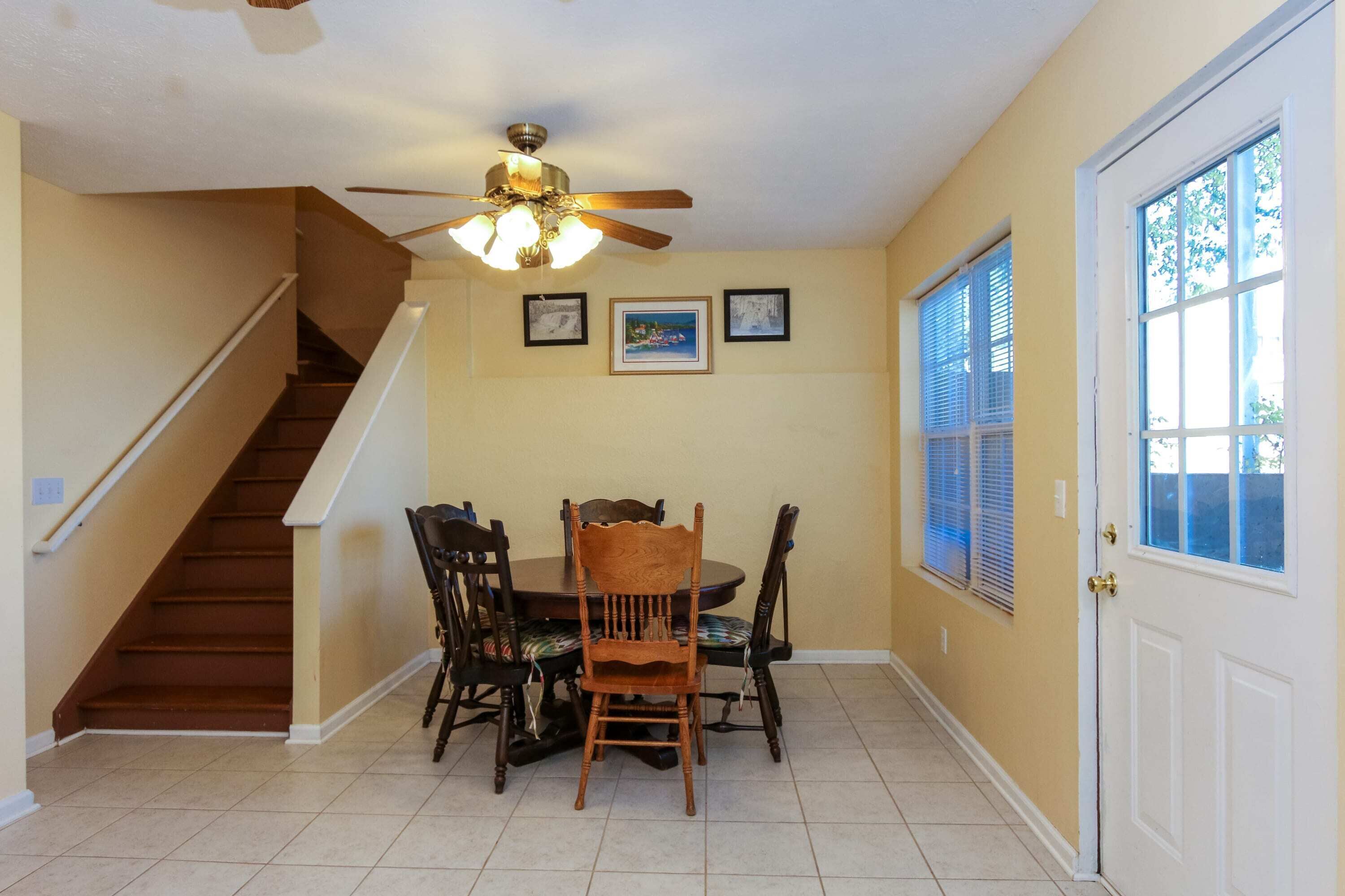 Un espace repas comprenant une table et des chaises en bois, un ventilateur de plafond, un escalier et une porte avec des fenêtres.