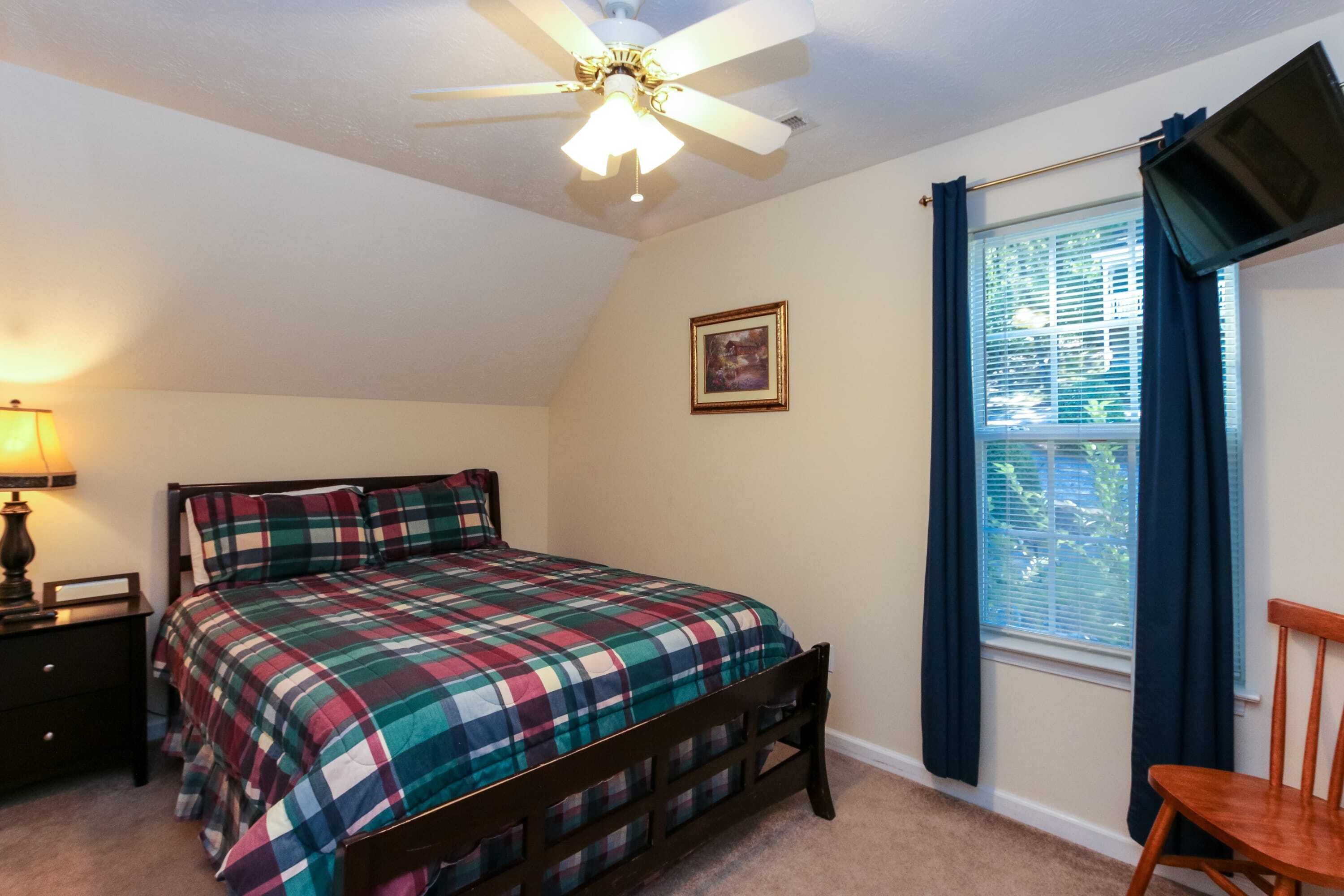 A bedroom with a bed, a ceiling fan, a nightstand, a chair, a window with curtains, and a framed picture on the wall.