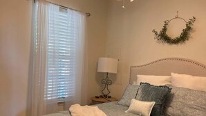 Ein Schlafzimmer mit einem Bett, einem Deckenventilator, einem Fenster mit Jalousien, einem Nachttisch mit Lampe und einem Wandkranz.
