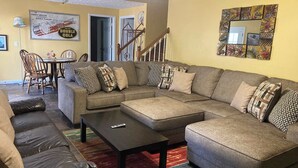 Ein Wohnzimmer mit Ecksofa, Couchtisch, Esstisch mit Stühlen, einer Treppe und einem wandmontierten Spiegel.