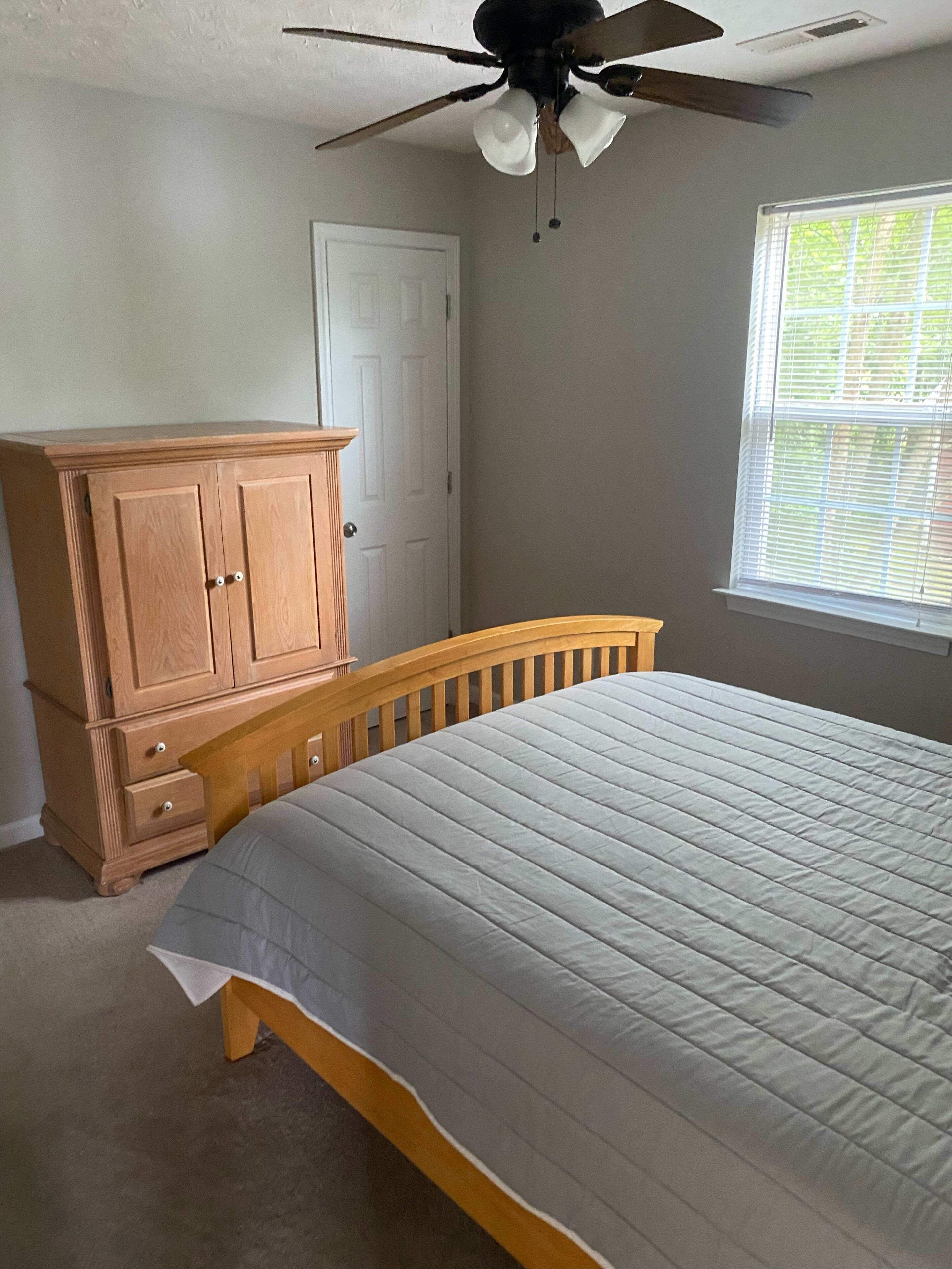 Une chambre à coucher comprenant un lit, une commode en bois, un ventilateur de plafond, une fenêtre avec des stores et une porte.