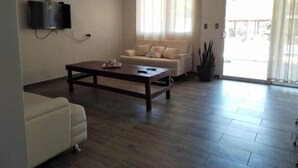 Ein Wohnzimmer mit einer Couch, einem Couchtisch, einem an der Wand montierten Fernseher und einer Glastür, die zu einem Außenbereich führt.