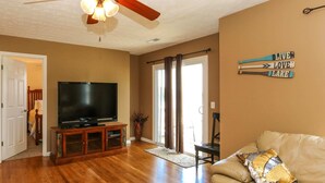 A living room with a wooden floor, a ceiling fan, a television on a wooden stand, a beige sofa, and a decorative sign on the wall. - House# 30 Endless View Not Pet-Friendly (Leesburg)