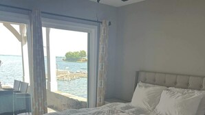 Un dormitorio con cama, ventilador de techo, vista al agua y una silla junto a la ventana.