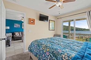 A bedroom with a bed, a ceiling fan, a TV, and a view of a lake.