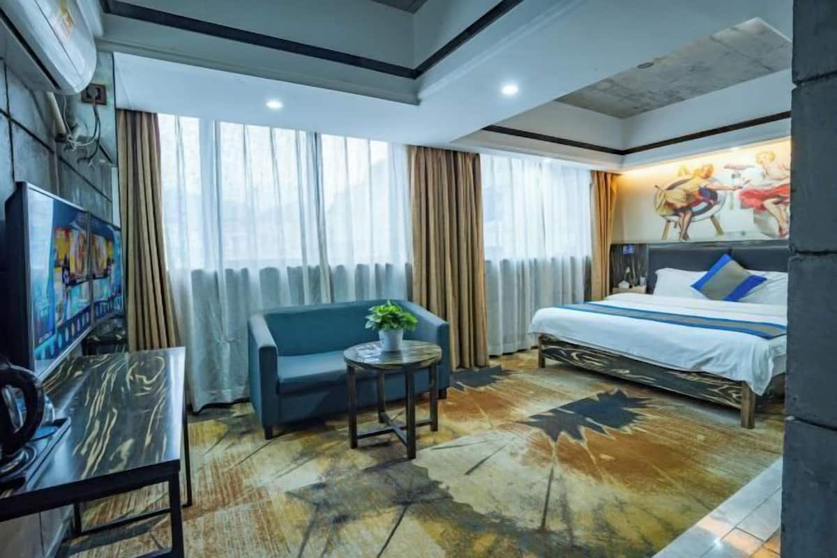 A hotel room with a bed, a sofa, a small table with a plant, a TV, and a large window with curtains.
