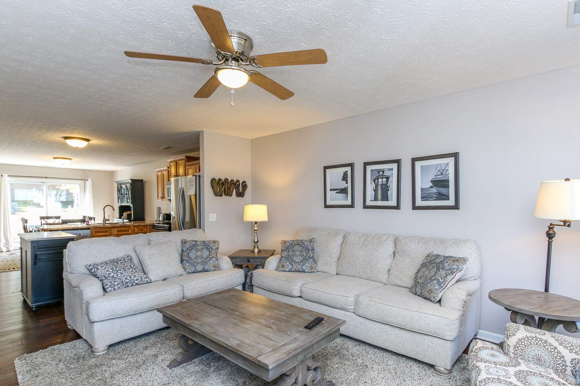 Un salon avec un canapé, une table basse et un ventilateur de plafond. On aperçoit une cuisine au fond, avec des placards et un réfrigérateur.