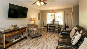 A living room with a flat-screen TV, a ceiling fan, a patterned armchair, a leather sofa, a dining table with chairs, and a window with curtains.