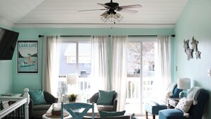 Una sala de estar con ventilador de techo, una mesa de comedor, sillas y un sofá. Hay una ventana con cortinas, un televisor montado en la pared y un cartel en la pared.