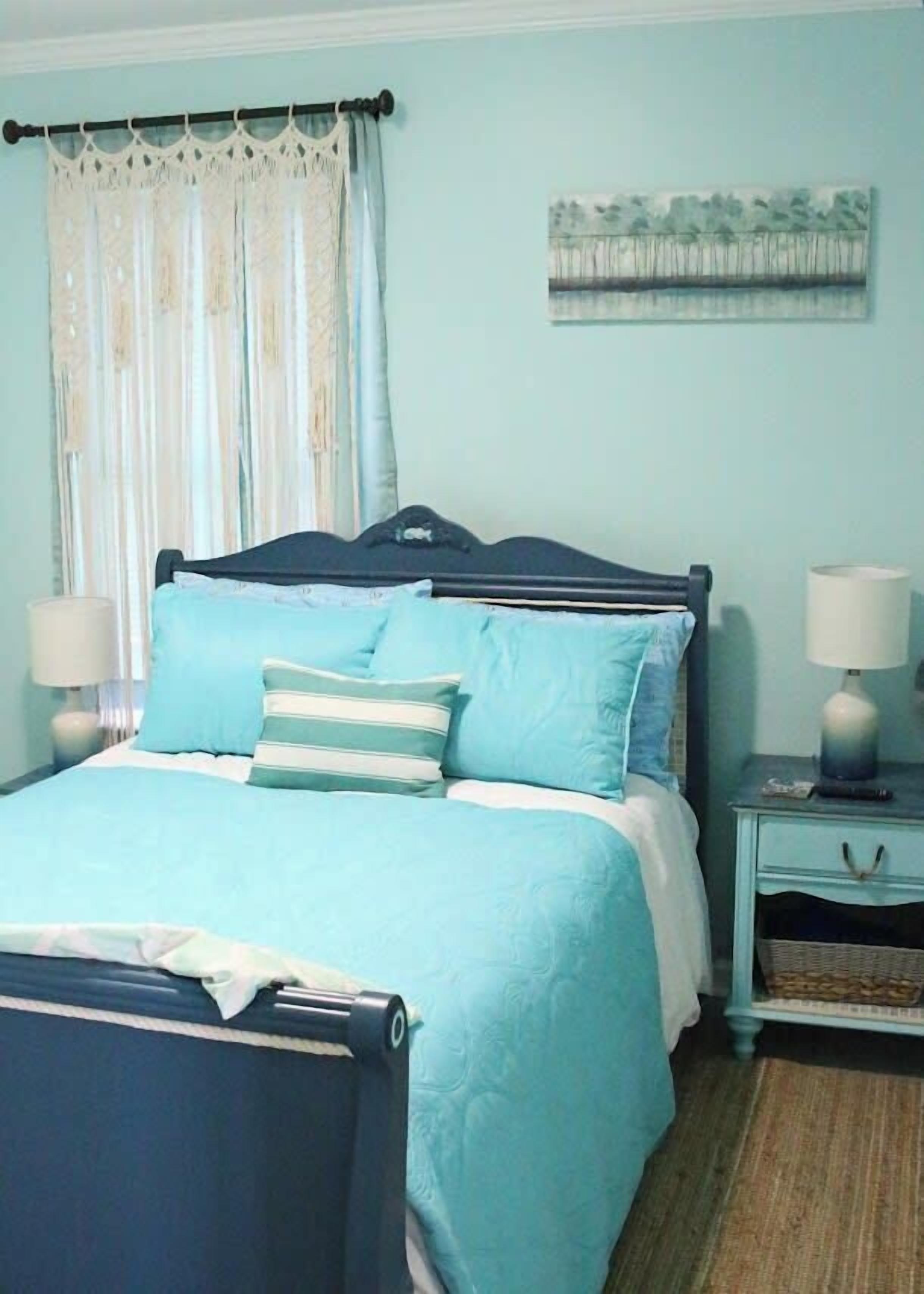 A bedroom with a bed, two bedside tables, a nightstand, a lamp, a curtain, and a painting on the wall.