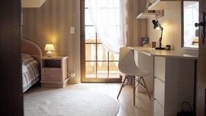 A bedroom with a bed, bedside table, lamp, and a desk with a chair. There is a window with curtains and a view outside.
