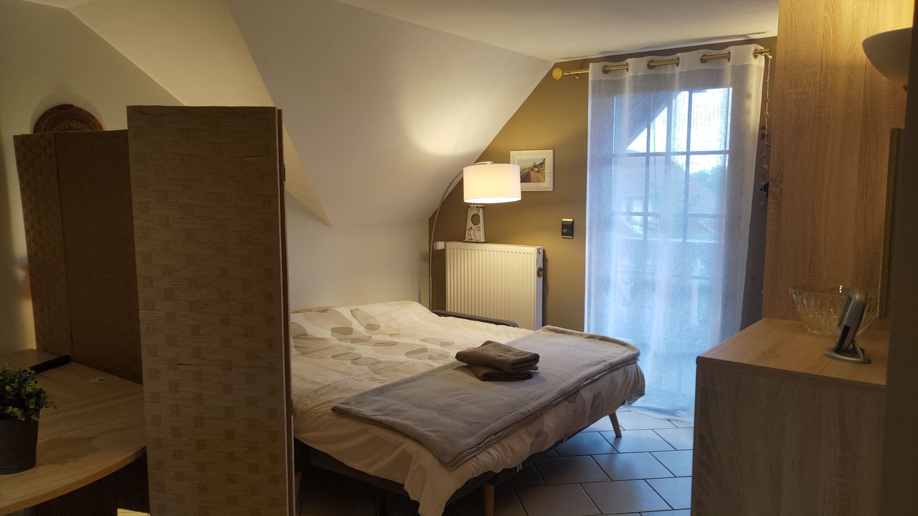 Une chambre à coucher avec un lit, une table de chevet, une lampe, un radiateur et une fenêtre avec des rideaux.